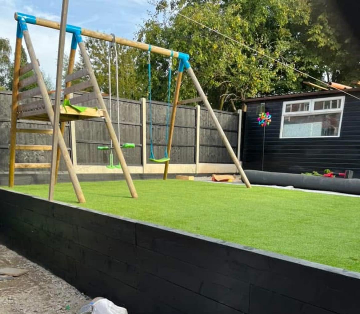 Raised artificial grass play area behind a black timber retaining wall, with a swing set