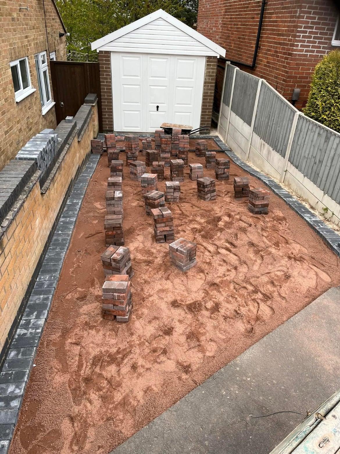 Red block paving packs set out across a sand laying course with charcoal edging in place