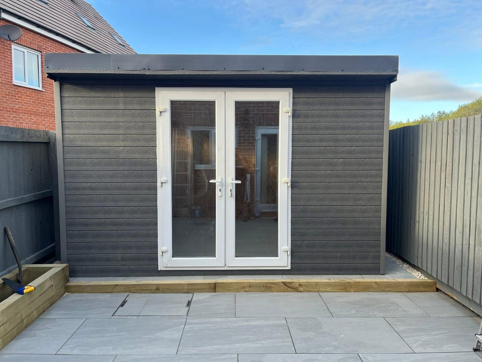 Grey clad garden room with French doors, fronted by a light porcelain patio and sleeper bed